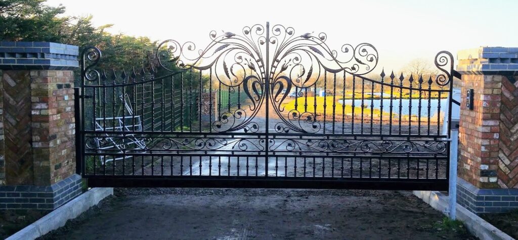Ornate black iron gate with scrollwork stands closed at a gated driveway, flanked by brick pillars.